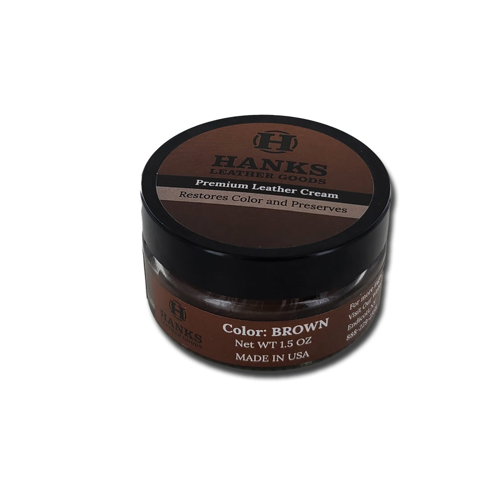 A round container of Hanks Belts Premium Leather Cream and Color Restoration with a brown label. Designed to restore and refinish leather, this premium leather cream preserves color. The container indicates the product color is brown, has a net weight of 1.5 ounces, and is made in the USA.