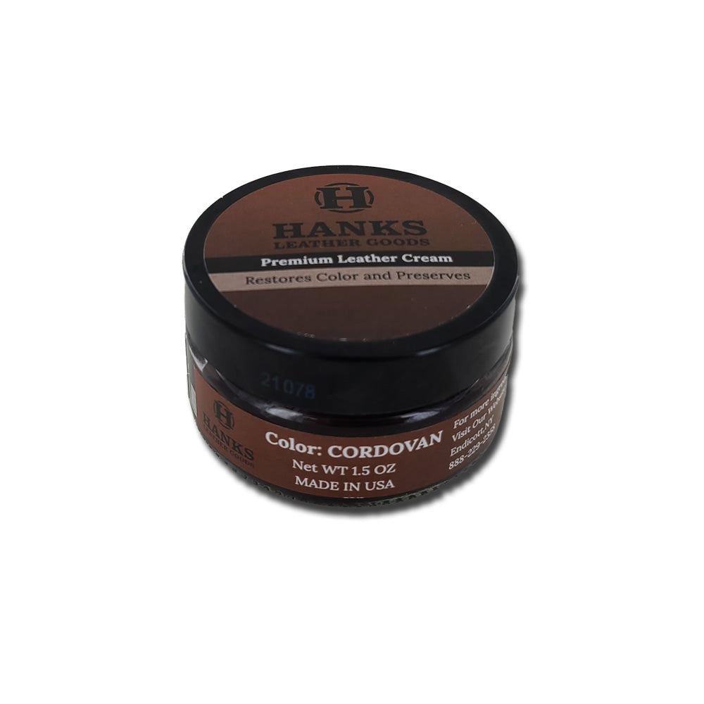 A small, round container of Hanks Belts' Premium Leather Cream and Color Restoration in Cordovan color. The label indicates the cream restores and refinishes leather products. The net weight is 1.5 ounces, and it is made in the USA. The container is black with a brown label.