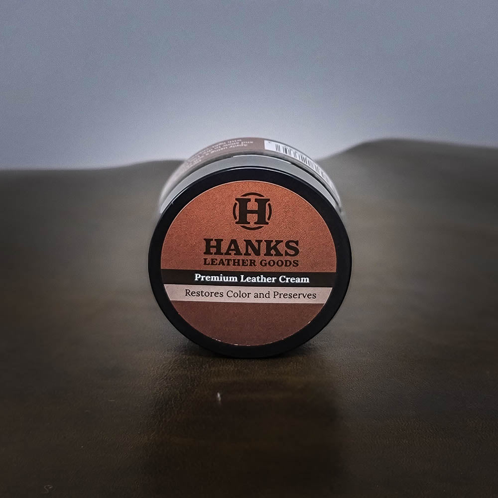 A small, round container of Hanks Belts Premium Leather Cream and Color Restoration is displayed on a dark surface. The label on the container reads "Hanks Belts," "Premium Leather Cream and Color Restoration," and "Restores Color and Preserves." Ideal for restoring and refinishing all your leather products.