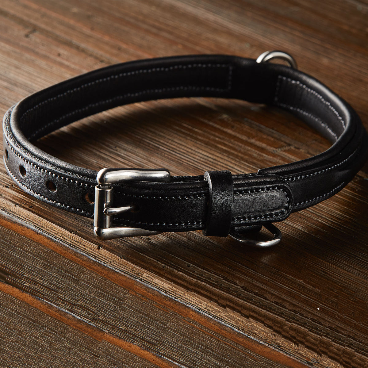 The Bentley Raised Collar by Hanks Belts, crafted from premium black English bridle leather with a silver buckle and stainless steel hardware, rests on a wooden surface.