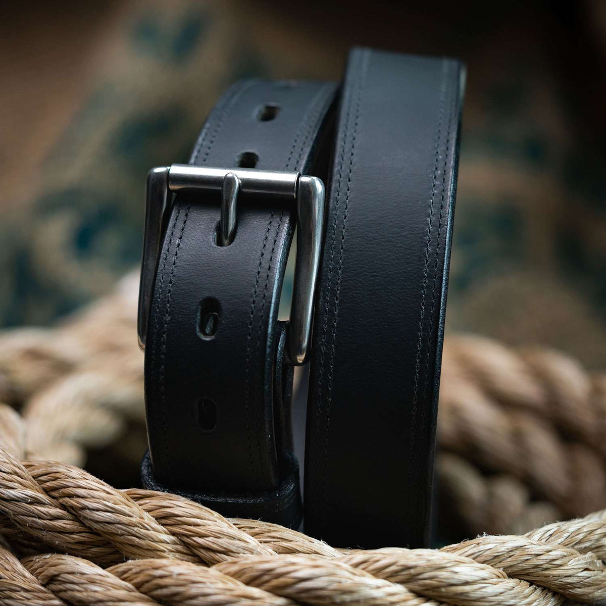 A close-up of Hanks Belts' The Montana Bison Double Layer, a rugged bison leather belt with a stainless steel buckle, resting on coiled tan rope against a blurred patterned background.