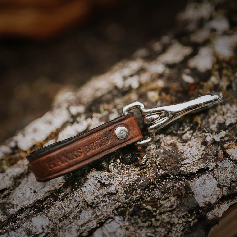 A leather Canyon Keychain - Keychain for Belts with a durable clip rests on a textured tree bark surface. The keychain is embossed with the text "Hanks Belts." The rich, brown leather contrasts with the rugged bark, creating a rustic and outdoorsy feel.