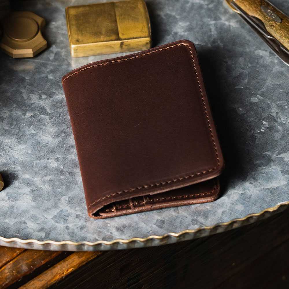 A closed Crater Bifold With Zipper from Hanks Belts, made of steer-hide leather, is placed on a round, metallic surface. Nearby, there is a brass lighter and the edge of a pocket knife is visible. The scene is warmly lit, creating a cozy, rustic ambiance.