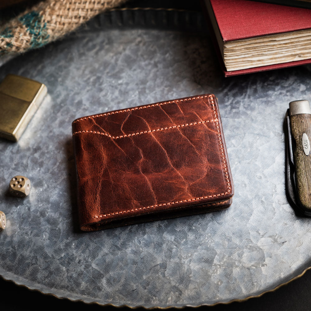 The Hanks Belts Powell Bison Bifold with ID Window Flap, crafted from bison leather with visible stitching, is showcased on a metal tray among a knife, dice, lighter, and books—perfect as a men's wallet or in a gift box.
