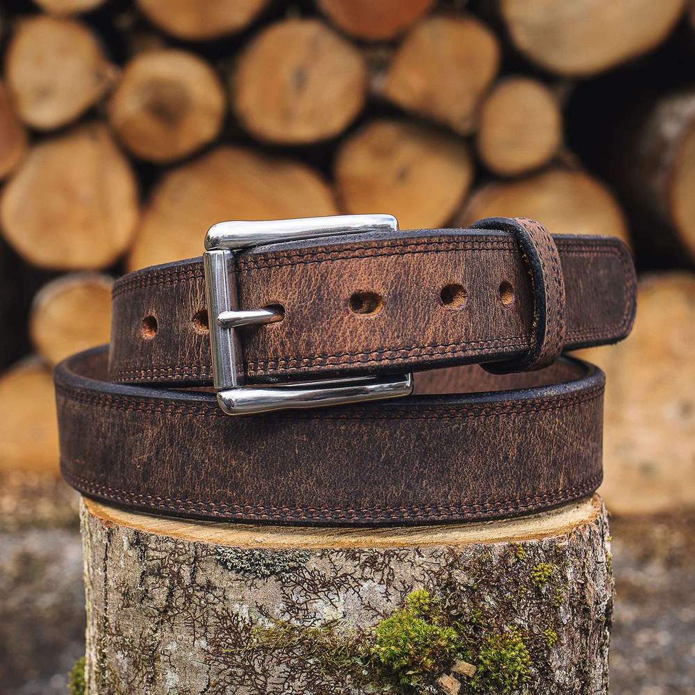 The Montana Bison Double Layer by Hanks Belts, featuring rugged bison leather and a stainless steel buckle, is coiled atop a mossy tree stump with blurred firewood logs in the background.