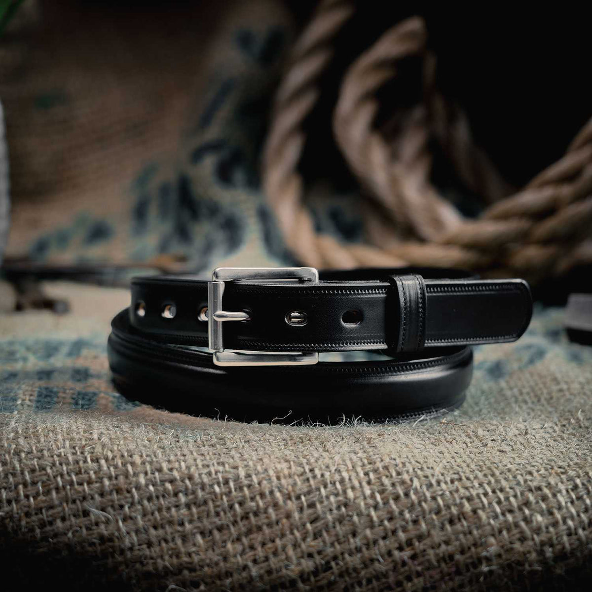 The Esquire Raised by Hanks Belts, a black English Bridle leather belt with a silver buckle, is coiled on textured burlap next to thick rope and a rustic, blurred backdrop.