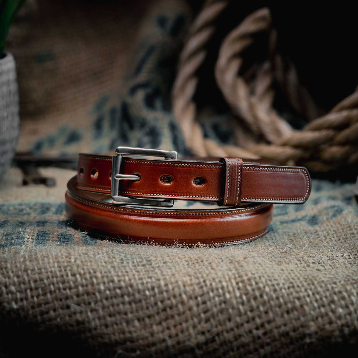 The Esquire Raised by Hanks Belts, made from rich English Bridle leather with a silver buckle, is coiled on burlap with rope and burlap fabric in the background.