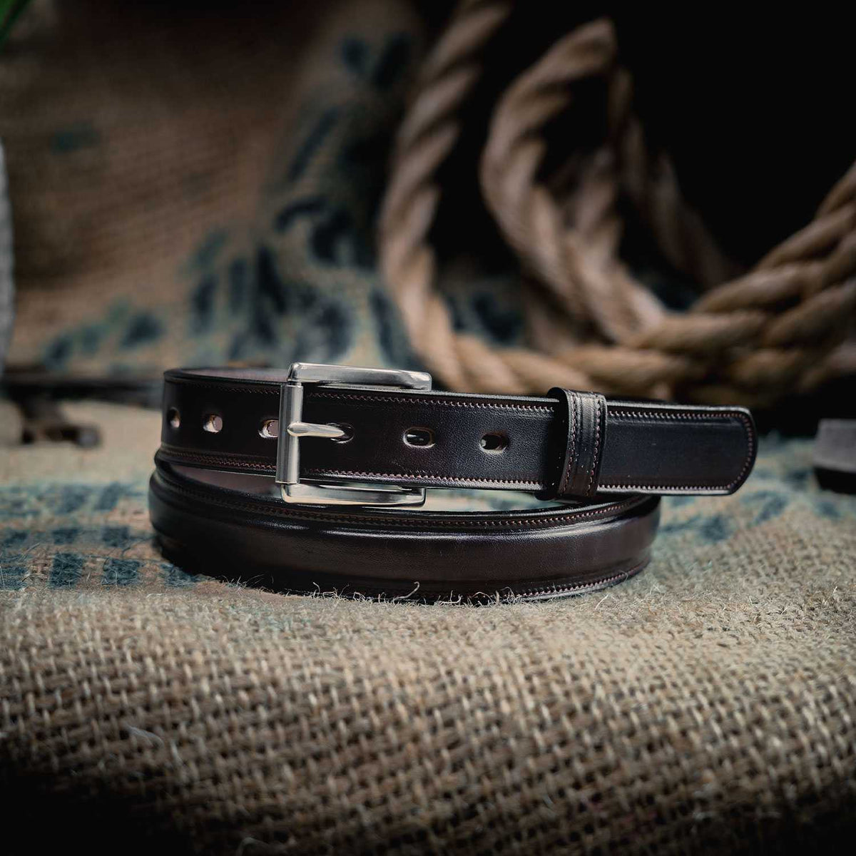 The Esquire Raised by Hanks Belts is a dark brown vegetable-tanned leather belt with white stitching and a silver buckle, coiled on textured burlap with a thick rope blurred in the background.