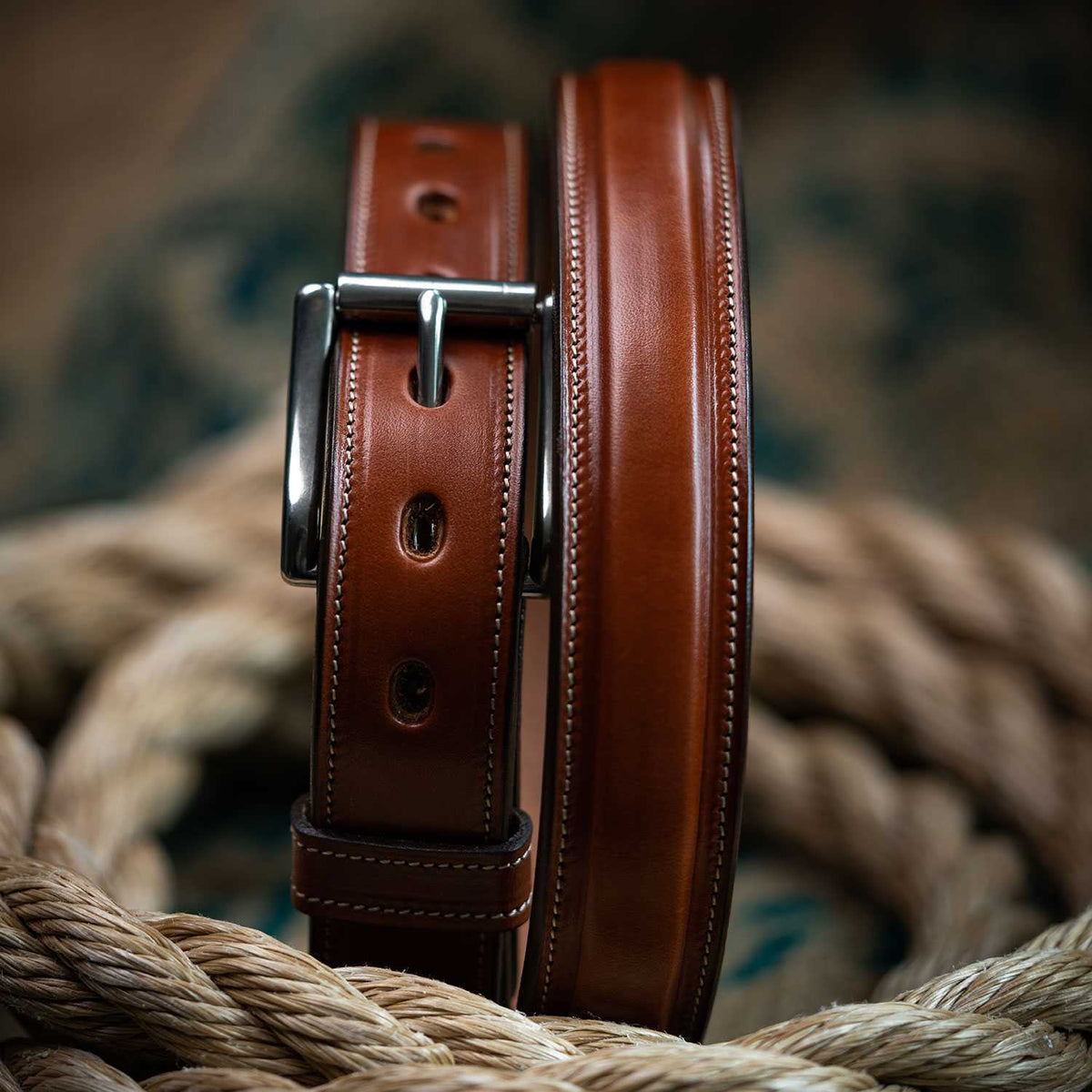 A close-up of The Esquire Raised by Hanks Belts, crafted from English Bridle leather with detailed stitching, rests on a thick coiled rope. Its silver buckle gleams against the rich brown leather and blurred background.