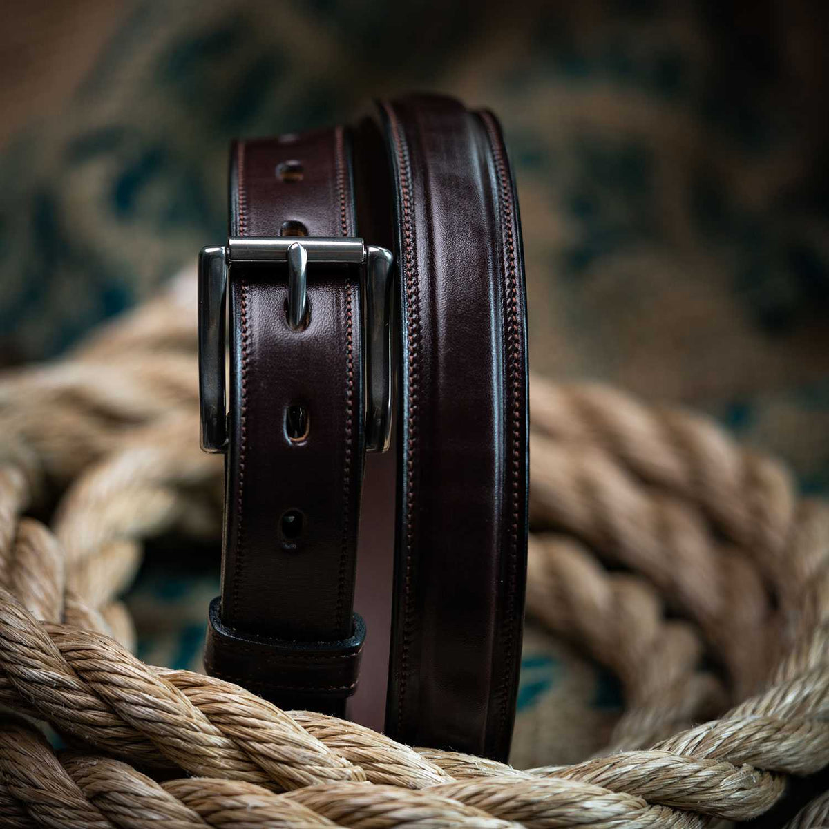 The Esquire Raised by Hanks Belts, a dark brown English Bridle leather belt with a silver buckle, is coiled upright and surrounded by thick, twisted rope on a textured surface.