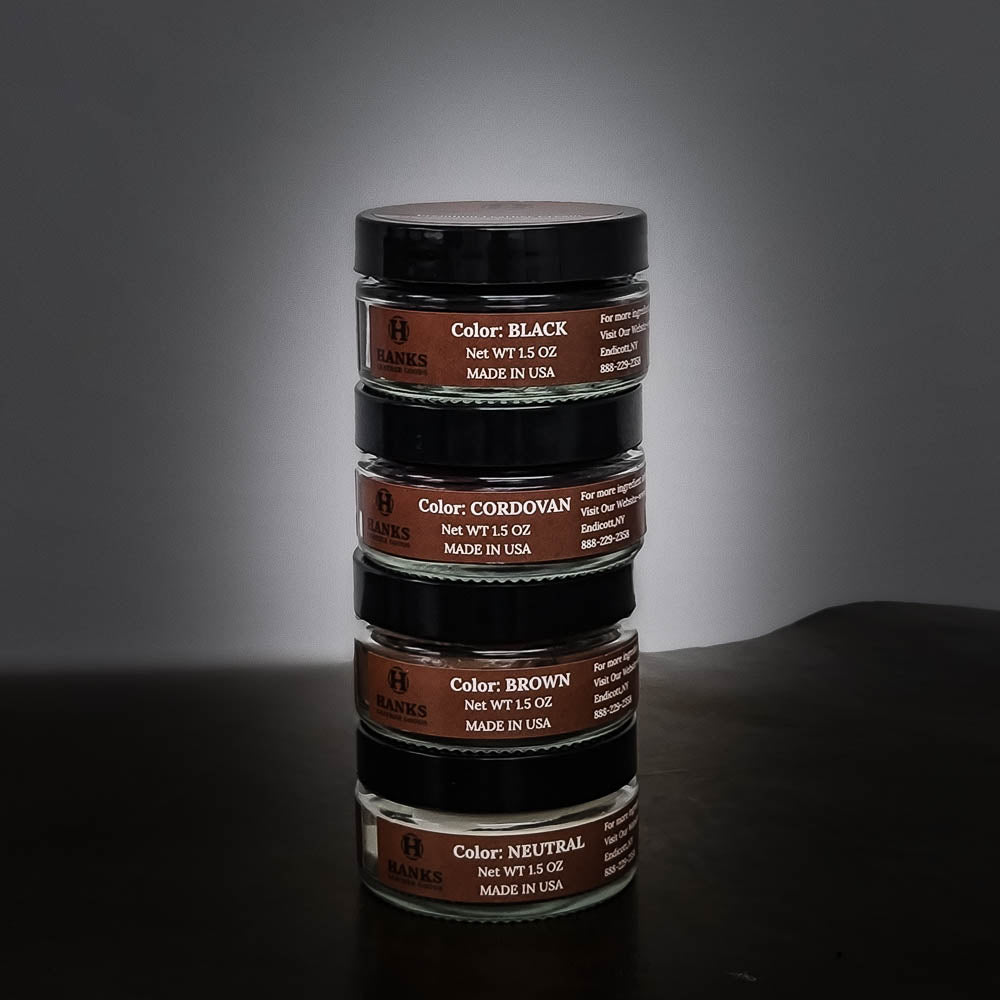 A stack of four containers of Hanks Belts' Premium Leather Cream and Color Restoration sits against a dark backdrop. Each container, perfect for restoring and refinishing leather products, lists its color and weight: Black (1.5 oz), Cordovan (1.5 oz), Brown (1.5 oz), and Neutral (1.5 oz).