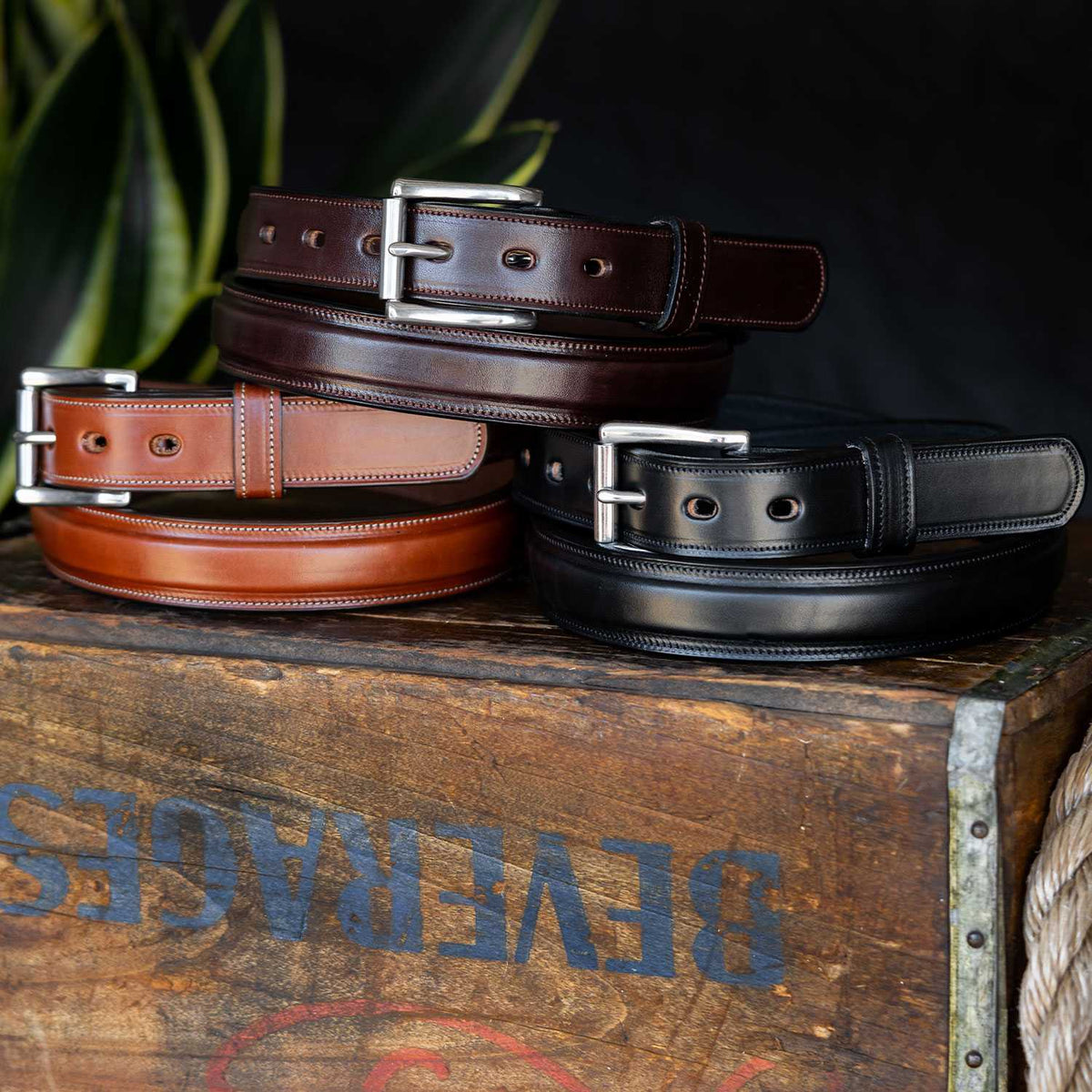 Three Esquire Raised belts by Hanks Belts—brown, tan, and black—made from premium vegetable-tanned leather, are neatly stacked on a rustic wooden box with faded lettering and green leaves in the background.