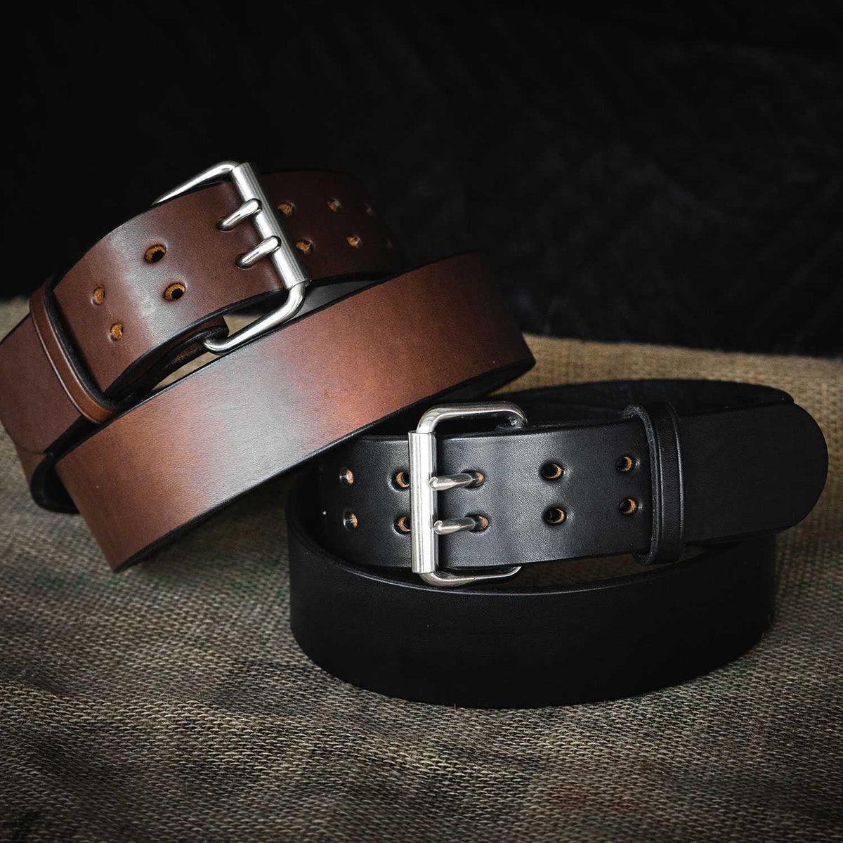 Two Hanks Belts “The Marshall” garrison style belts, one brown and one black with silver buckles, are coiled and overlapping on a textured burlap surface, showcasing their sturdy build and smooth finish.