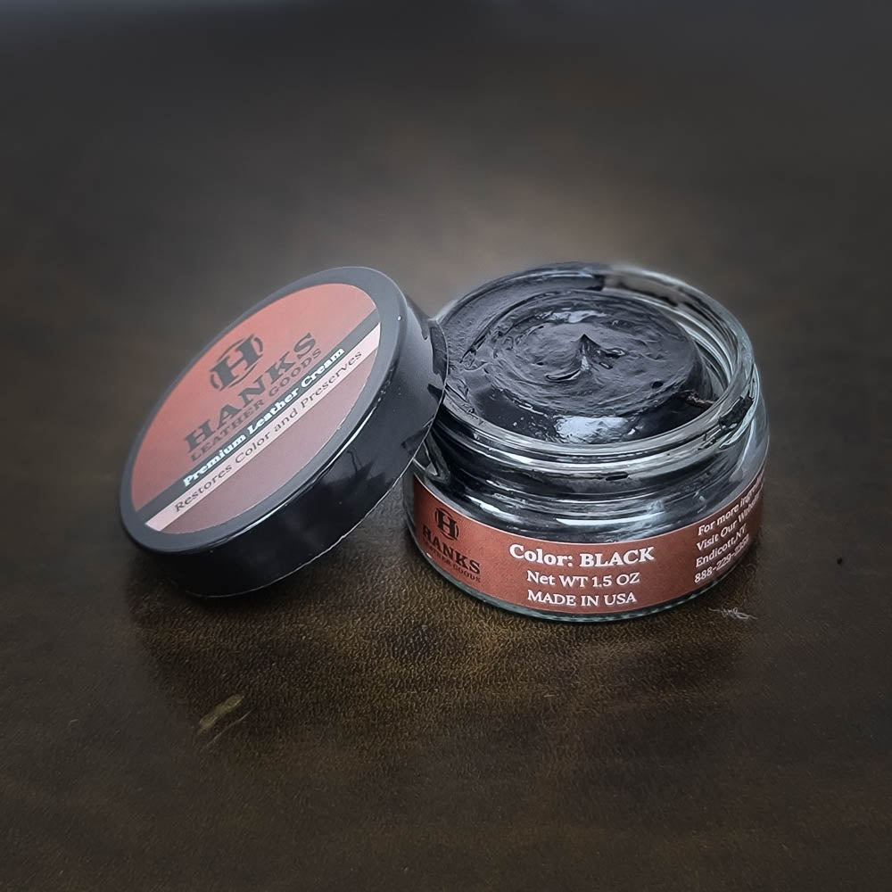 A small glass jar of Hanks Belts Premium Leather Cream and Color Restoration is shown with the lid off and leaning against the jar. The label features a logo, description, and the text "Color: BLACK Net WT 1.5 OZ MADE IN USA" on a dark background.