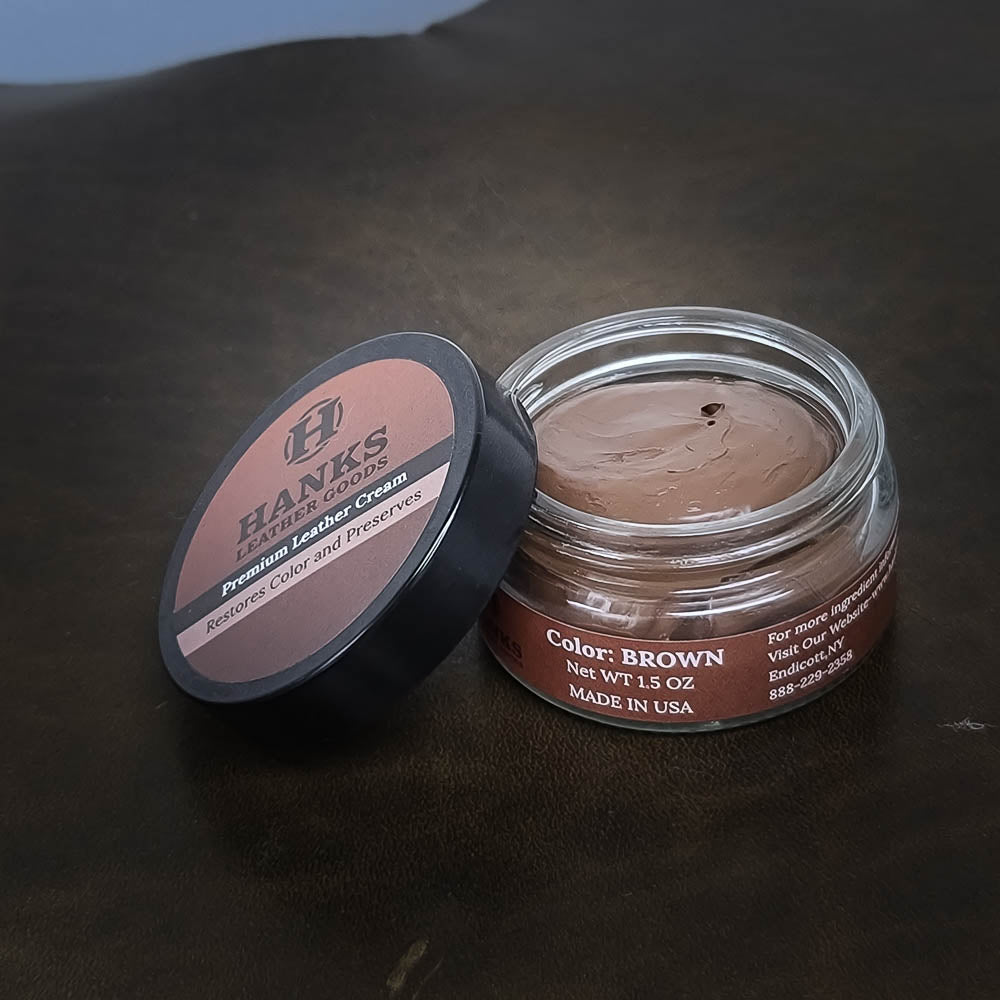 A small, open jar of Hanks Belts Premium Leather Cream and Color Restoration sits on a dark surface, its lid placed beside it. The label reads "Brown," net weight 1.5 ounces, and "Made in USA." This premium leather care product is perfect for maintaining Hanks Belts and other fine leather goods.
