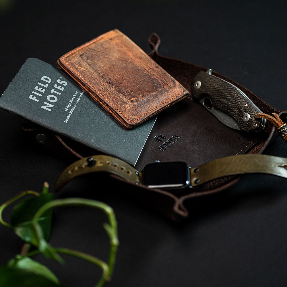 A leather pouch lies open on a dark surface, revealing a "FIELD NOTES" notebook, a brown cardholder, and a folding knife. Next to it is a smartwatch with a brown leather strap. A Hanks Belts Premium Leather Valet Tray holds everything in place, and a green plant is partially visible in the corner.