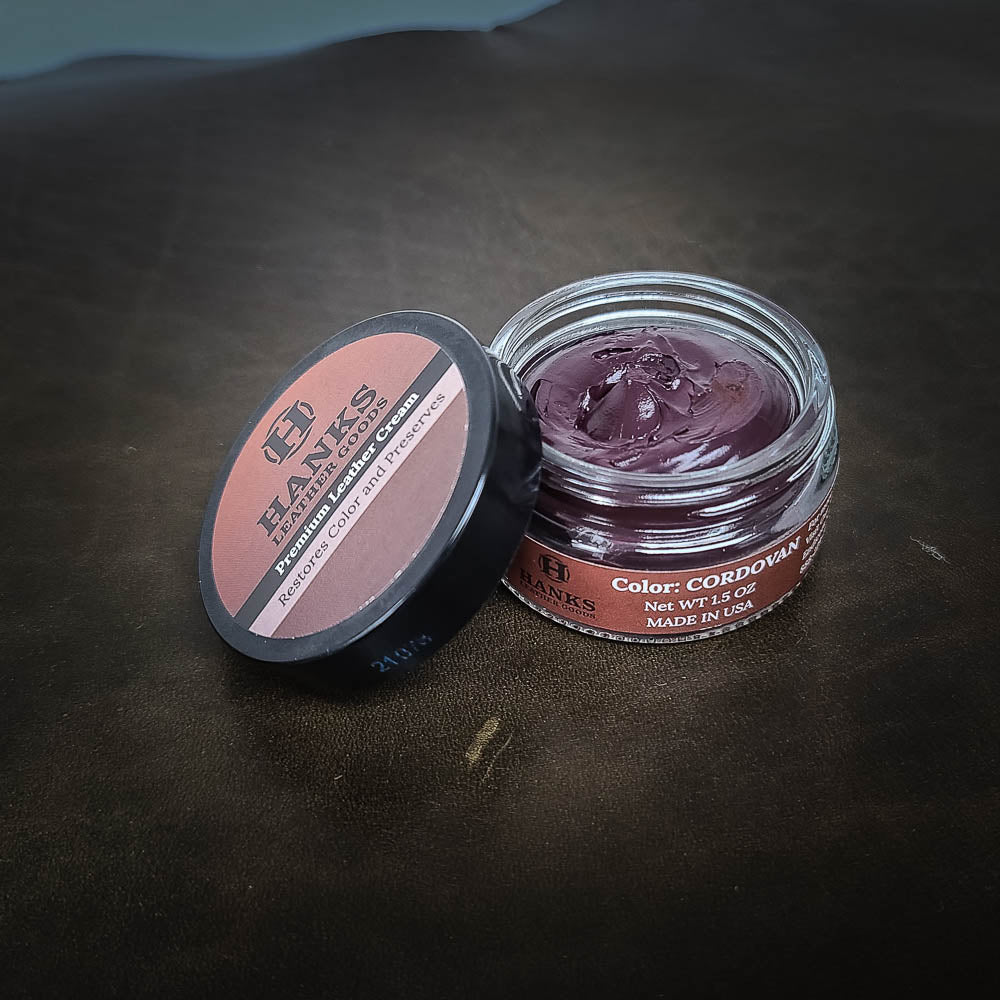 A small, open jar of Hanks Belts Premium Leather Cream and Color Restoration, color Cordovan, sits on a dark surface. The translucent leather cream inside is deep burgundy. The lid, placed next to the jar, displays the brand name and product details. Ideal for premium leather care alongside your Hanks Belt.
