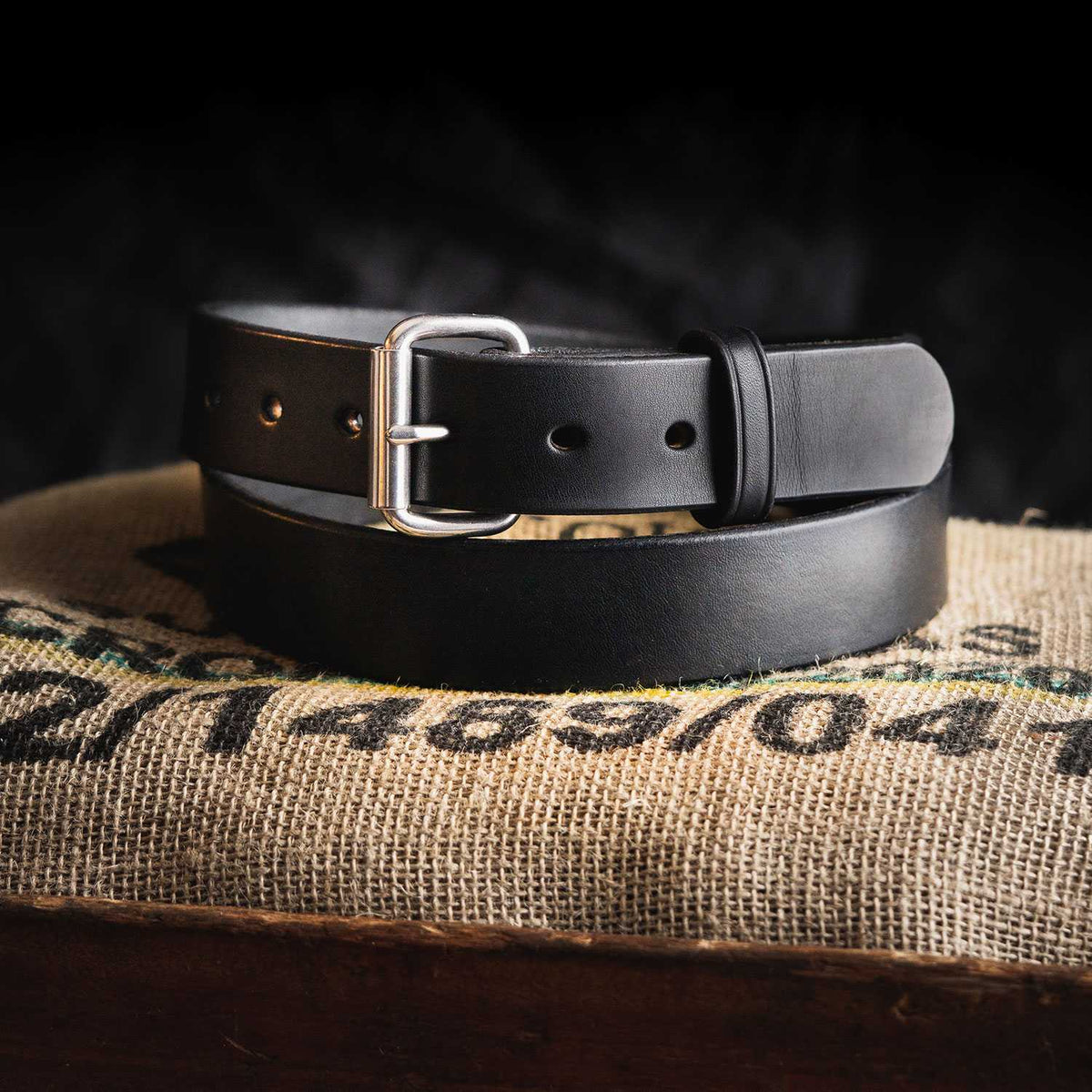 The Gunner by Hanks Belts, a black full-grain steer hide belt with a silver buckle, is coiled on a burlap sack with printed numbers, set against a dark, blurred background.