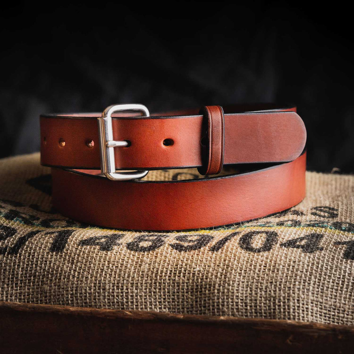 The Gunner by Hanks Belts—a brown full-grain steer hide belt with a silver buckle—is coiled on a burlap sack marked with black and green text, set against a dark background.