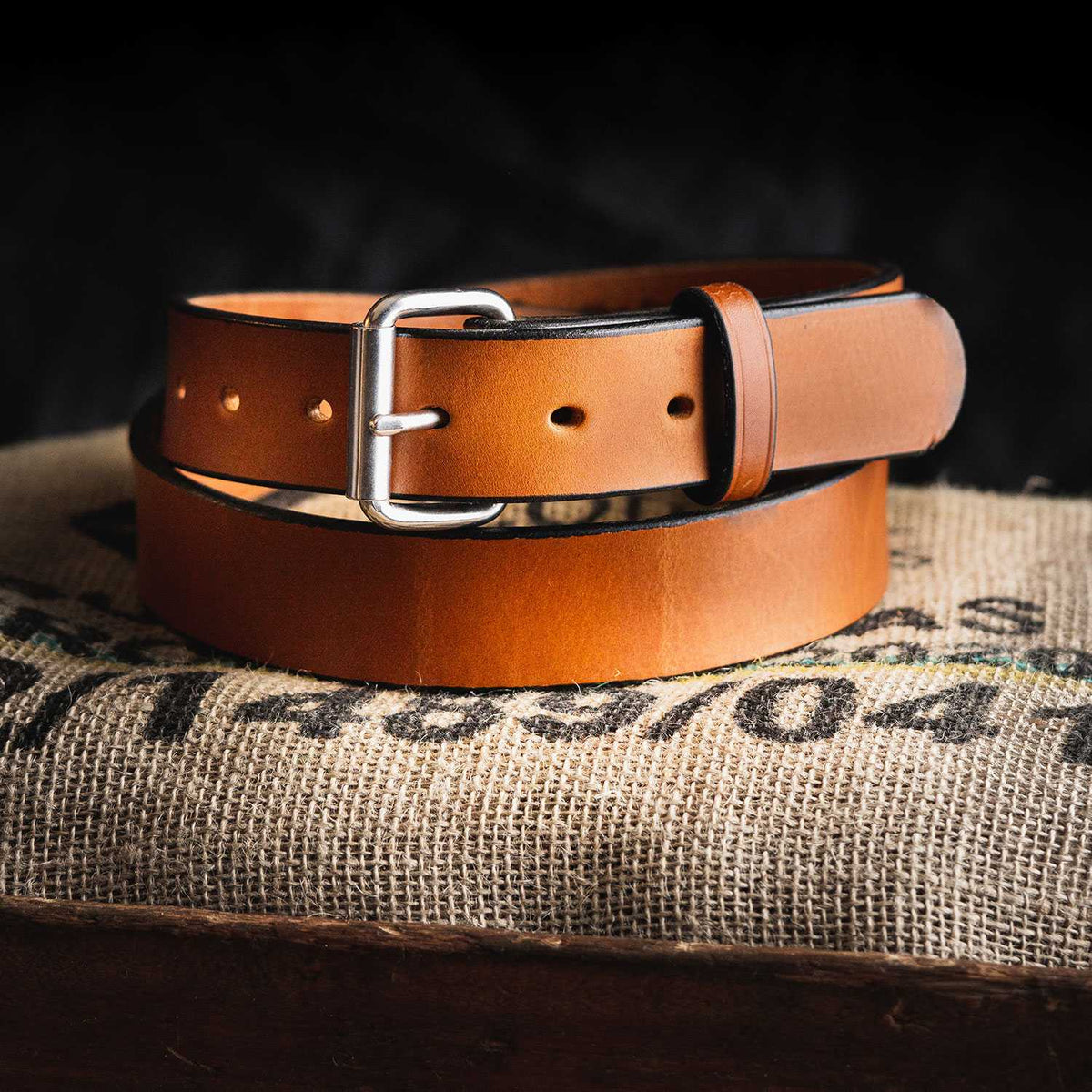 The Gunner by Hanks Belts, a tan full-grain steer hide belt with a silver buckle, is coiled on a burlap sack against a dark background.