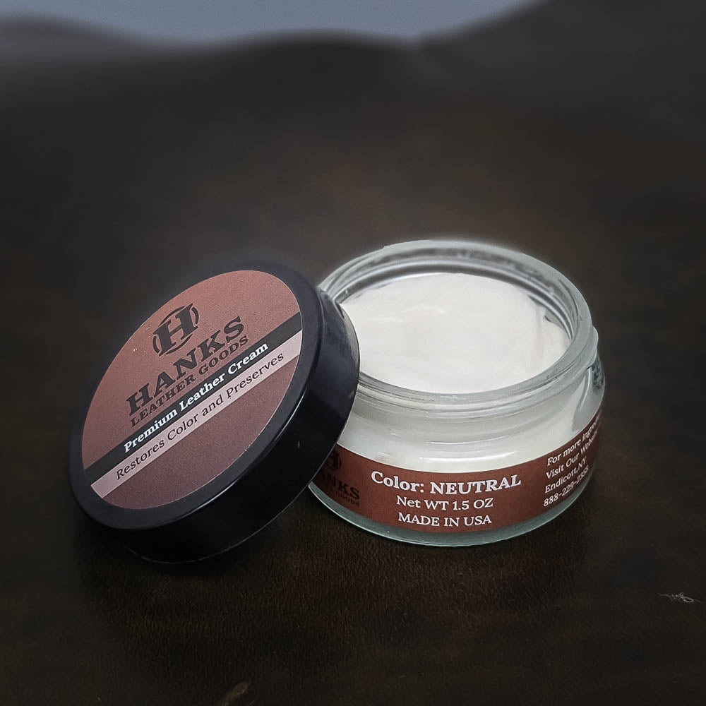 A small open jar of Hanks Belts Premium Leather Cream and Color Restoration is shown, promising to "Restore and Refinish" with its neutral-colored formula. Perfect for use on your favorite Hanks Belt, this 1.5 oz cream works wonders in restoring color and preserving your leather goods. The lid sits partially atop the jar, ready for your next use.