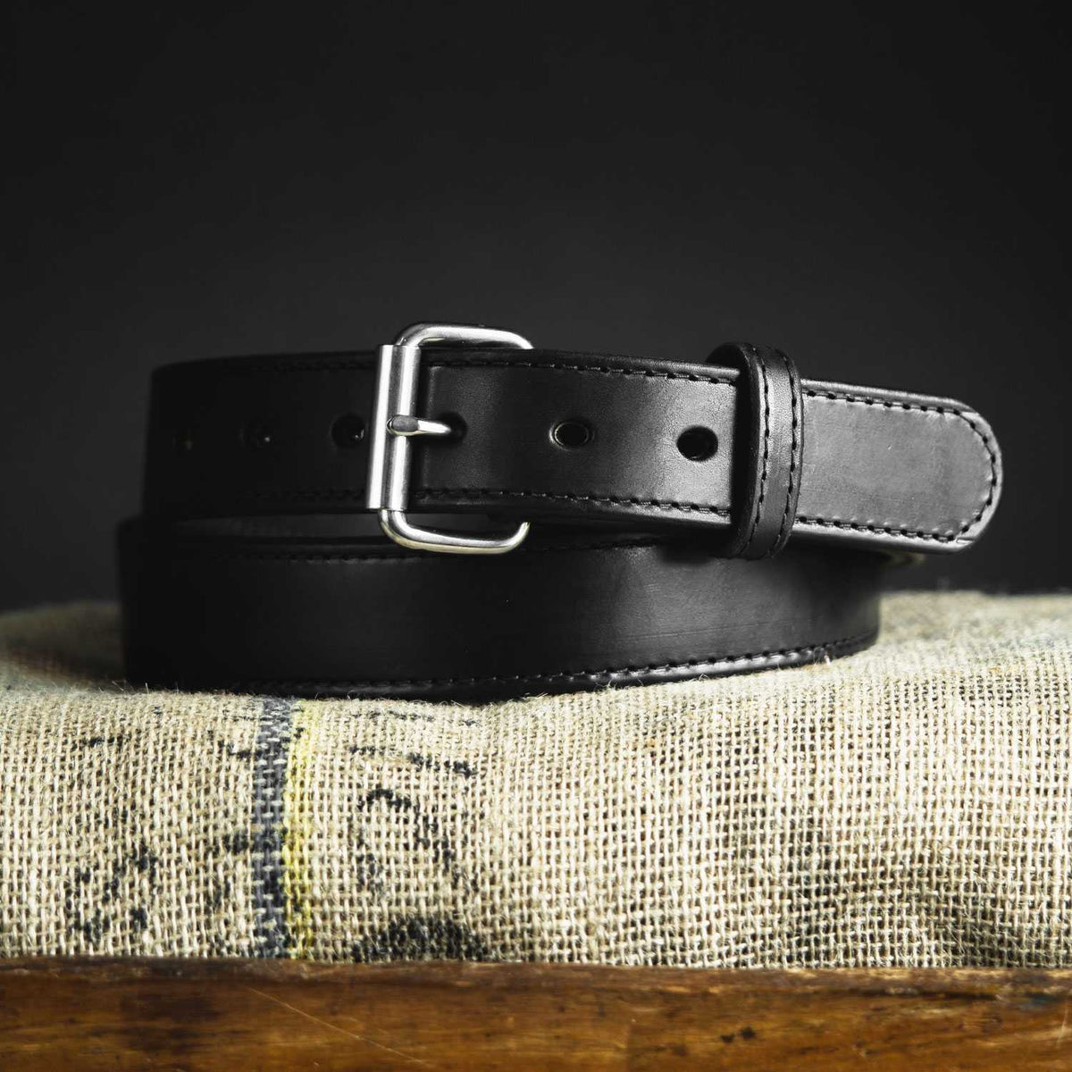 A sleek Hanks Belts Steel Core Belt - 1.5" in black leather with a silver buckle is neatly coiled and placed on a piece of burlap cloth against a dark background.