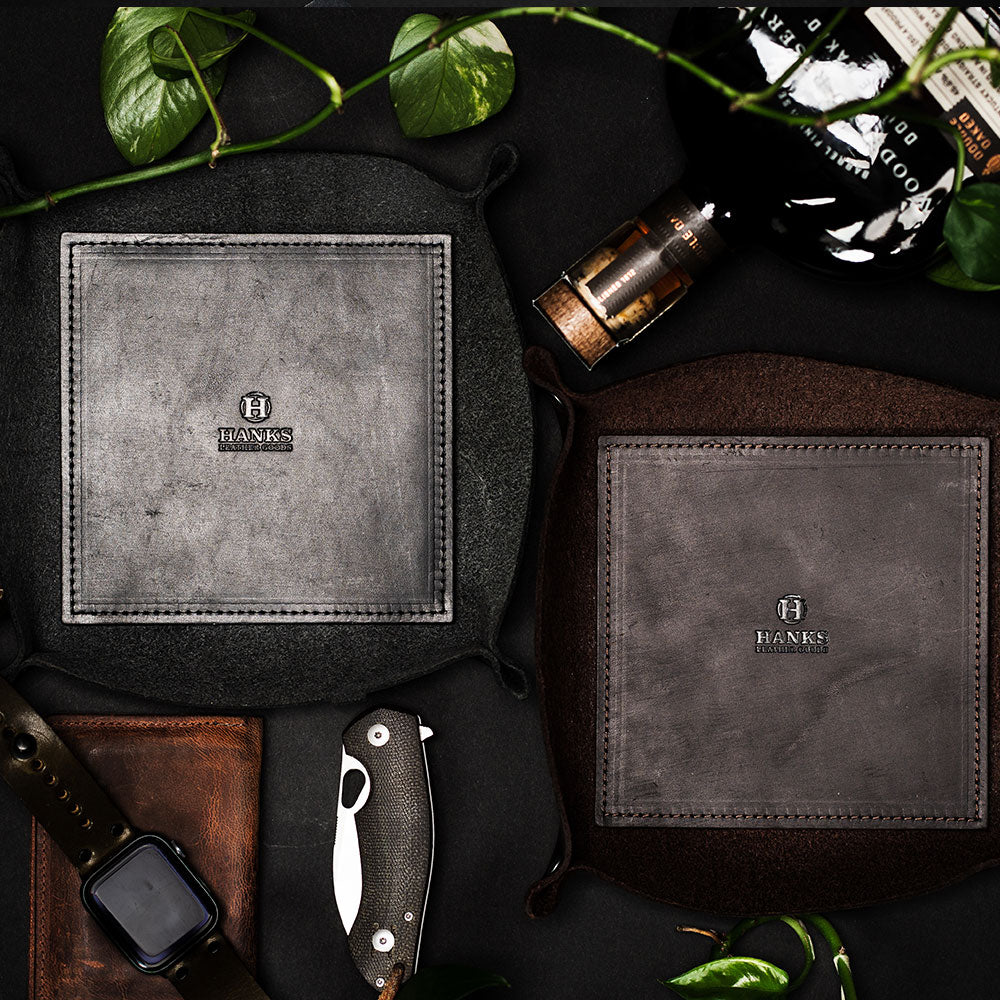 Two Premium Leather Valet Trays by Hanks Belts are placed on a dark surface. Surrounding them are an Apple Watch, a folding knife, a brown belt, a small bottle, and green plant leaves. The USA-made trays in gray and dark brown colors provide a rustic and organized aesthetic.