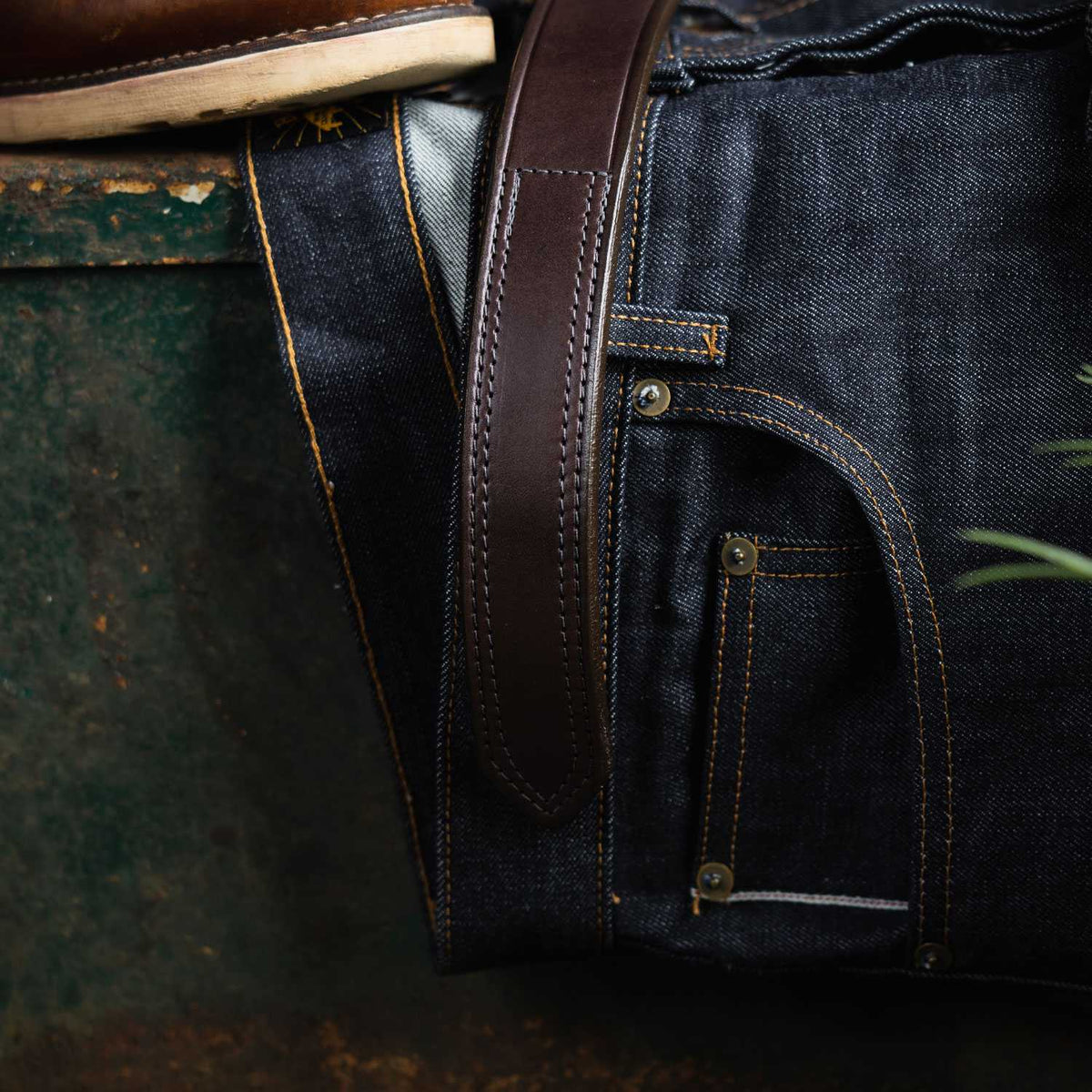 A close-up shows dark blue denim jeans with orange stitching, Hanks Belts' The Scratchless Mechanics full grain leather belt threaded through the loops, and part of a brown shoe visible near the top left.