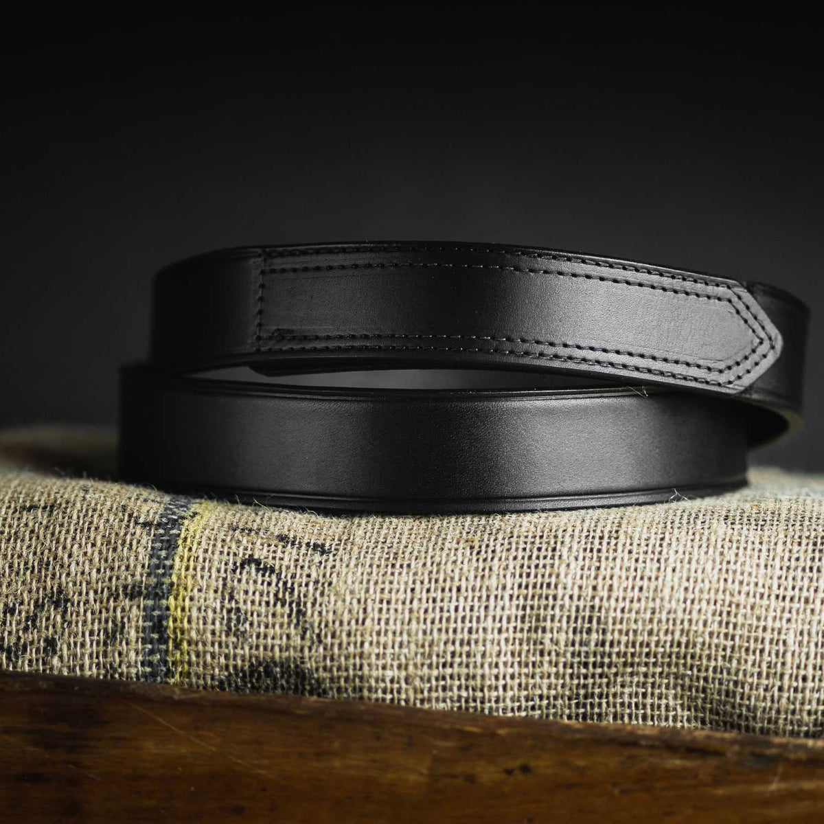 A close-up of The Scratchless Mechanics by Hanks Belts, a genuine full grain leather belt with detailed stitching, coiled on burlap against a dark background.
