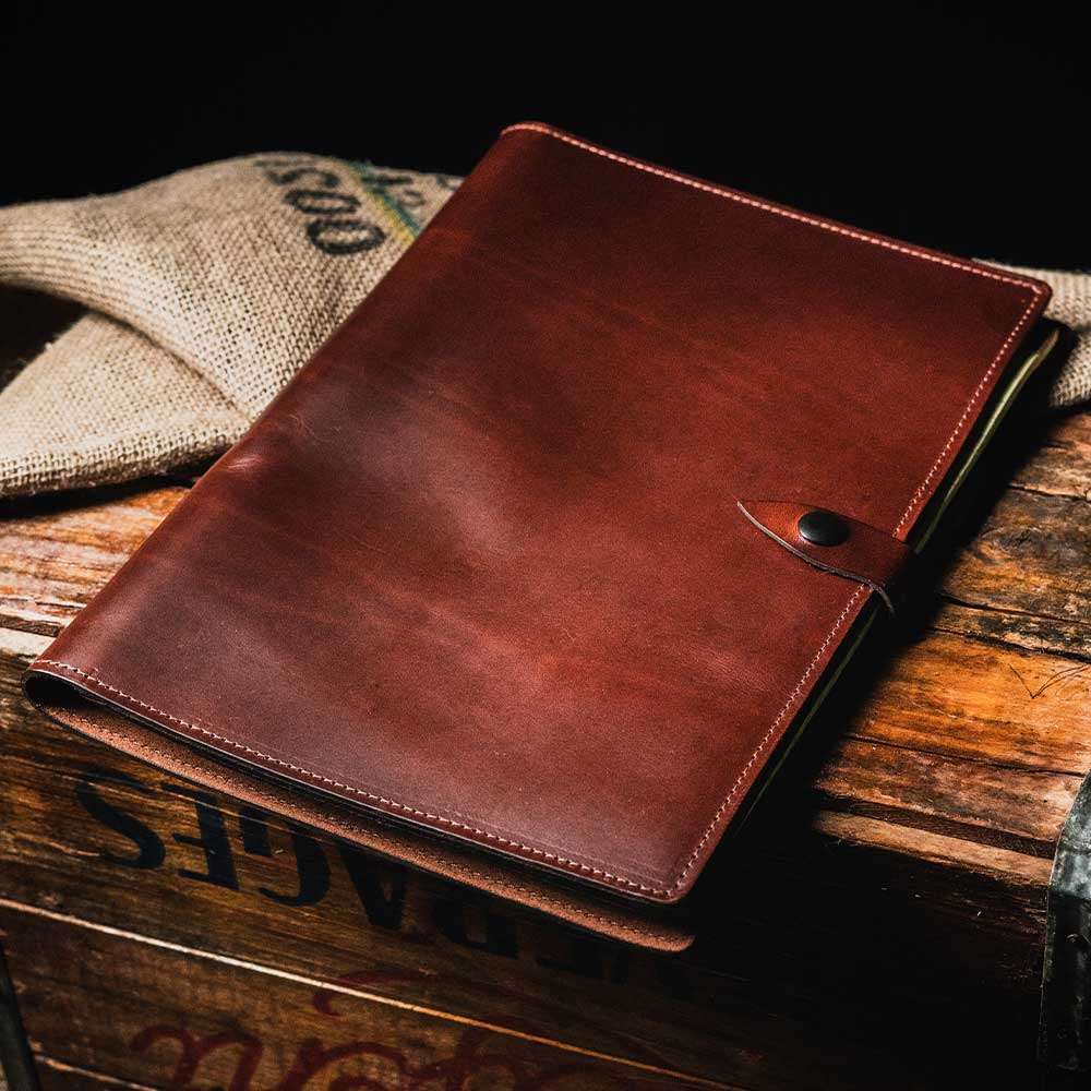 The Hanks Belts Luxury Folio, a brown, USA-made leather notebook with a button closure, sits on a wooden crate with a burlap sack partially in view. The dark setting gives the scene a rustic and vintage feel, highlighting the full-grain leather's texture and craftsmanship.