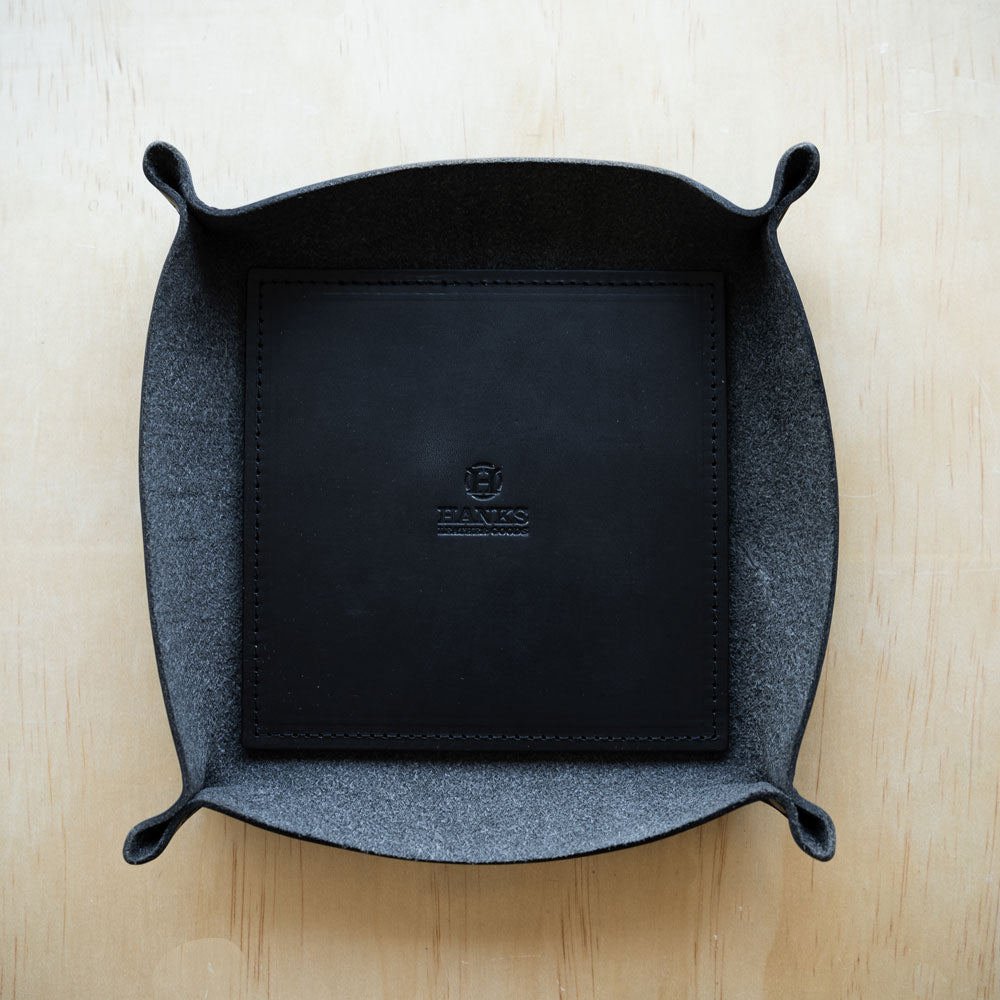 A stylish Hanks Belts Premium Leather Valet Tray with raised corners, featuring the embossed logo "Hanks" at the bottom center, rests on a light wooden surface. The interior is lined with a soft gray material, creating a striking contrast with the black exterior.