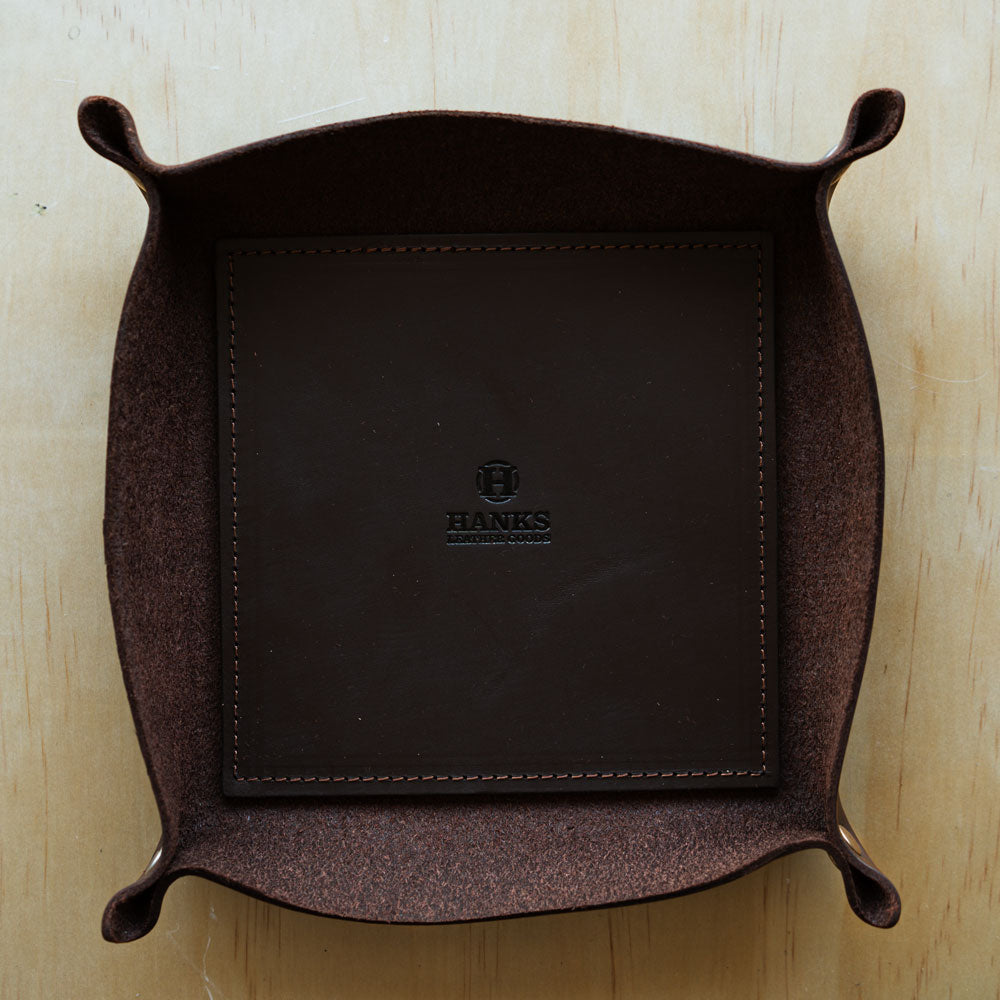 A Premium Leather Valet Tray from Hanks Belts, made in the USA, sits on a wooden surface. The tray is crafted from brown full-grain leather with pinched bridle leather corners for a structured shape. Its interior base is dark brown and features an embossed logo in the center with the name "Hanks" beneath it.