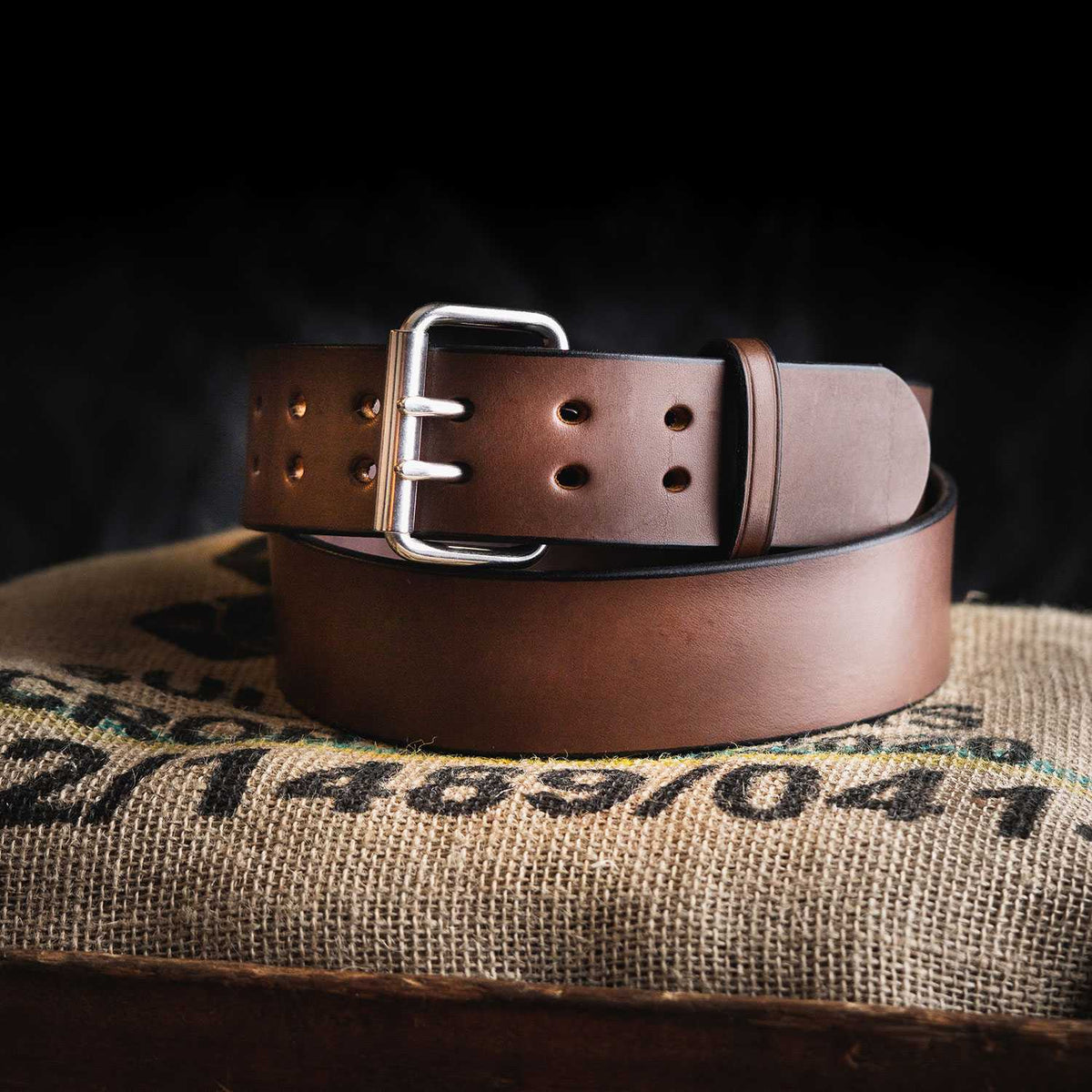 The Marshall by Hanks Belts, a brown leather CCW gun belt with a silver buckle, is coiled neatly on a textured burlap sack against a dark background.