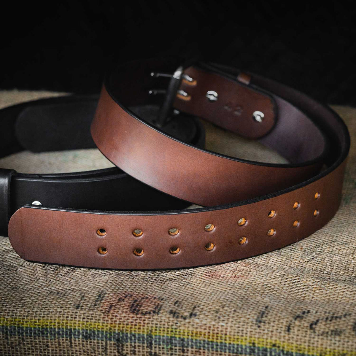 A close-up of The Marshall by Hanks Belts, one brown and one black leather belt with metal buckles on textured fabric—ideal for weightlifting or as a durable CCW Gun Belt for concealed carry.
