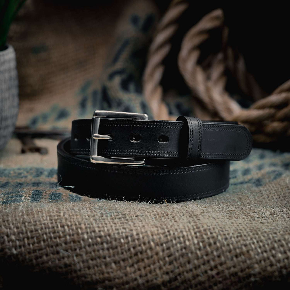 The Montana Bison Double Layer by Hanks Belts, made of rugged bison leather with a stainless steel buckle, is coiled on burlap with a thick rope and textured cloth blurred in the background.