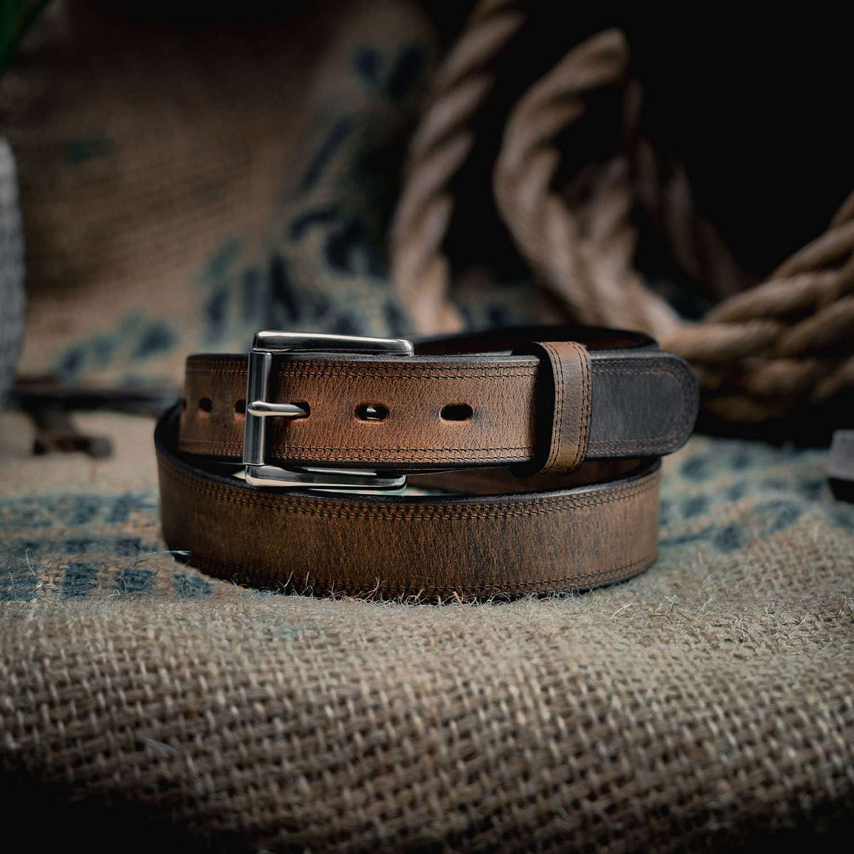 The Montana Bison Double Layer by Hanks Belts, featuring rugged bison leather and a stainless steel buckle, is coiled on burlap with a thick rope and rustic background elements.