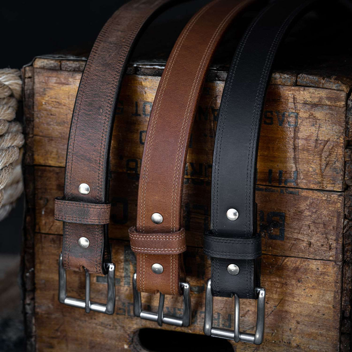 Three Hanks Belts Montana Bison Double Layer belts—dark brown, light brown, and black—are displayed on a rustic crate. Each features rugged leather, a stainless steel buckle, and metal rivets for sturdy style.