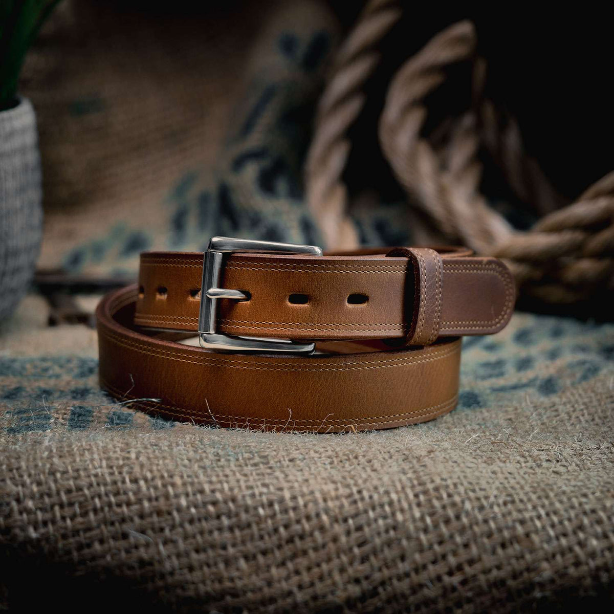 The Montana Bison Double Layer belt by Hanks Belts, crafted from rugged bison leather with a silver buckle, is coiled on a textured burlap surface. A thick rope and a plant pot are softly blurred in the background.