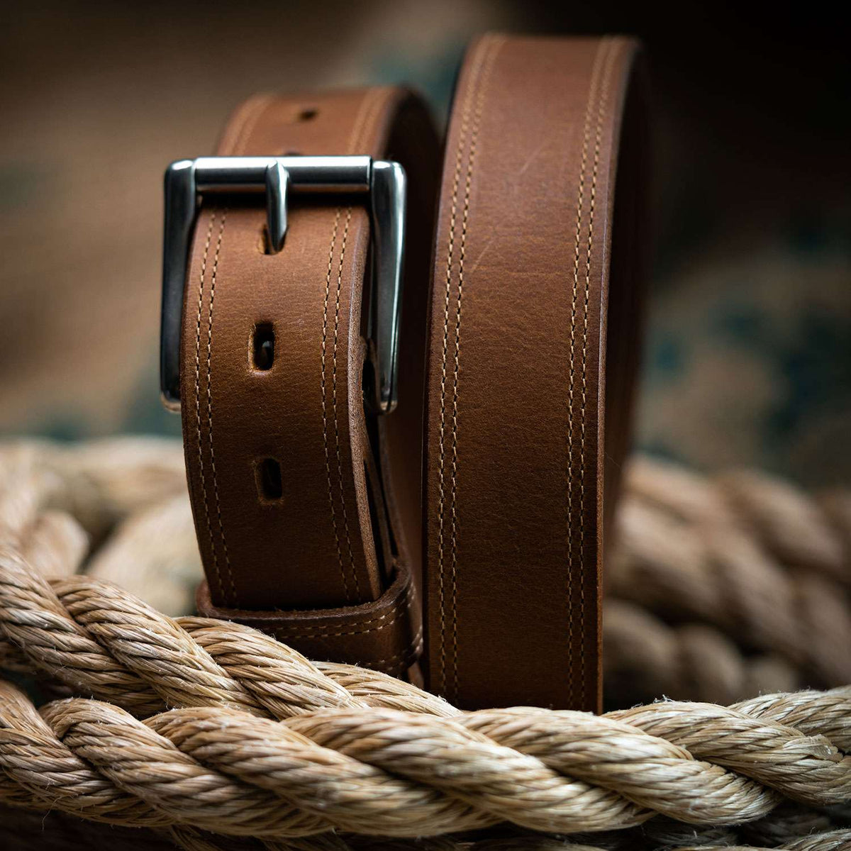 The Montana Bison Double Layer belt by Hanks Belts, crafted from rugged bison leather with white stitching and a stainless steel buckle, is coiled atop thick beige rope against a softly blurred background.