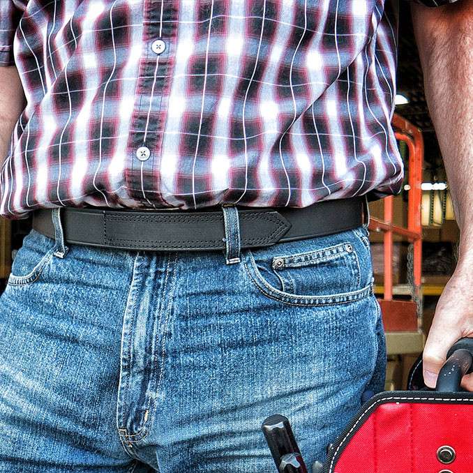 A person in a red plaid shirt and blue jeans holds a red tool bag, with the image focused on the torso, one arm, and The Scratchless Mechanics full grain leather belt by Hanks Belts worn around the waist.