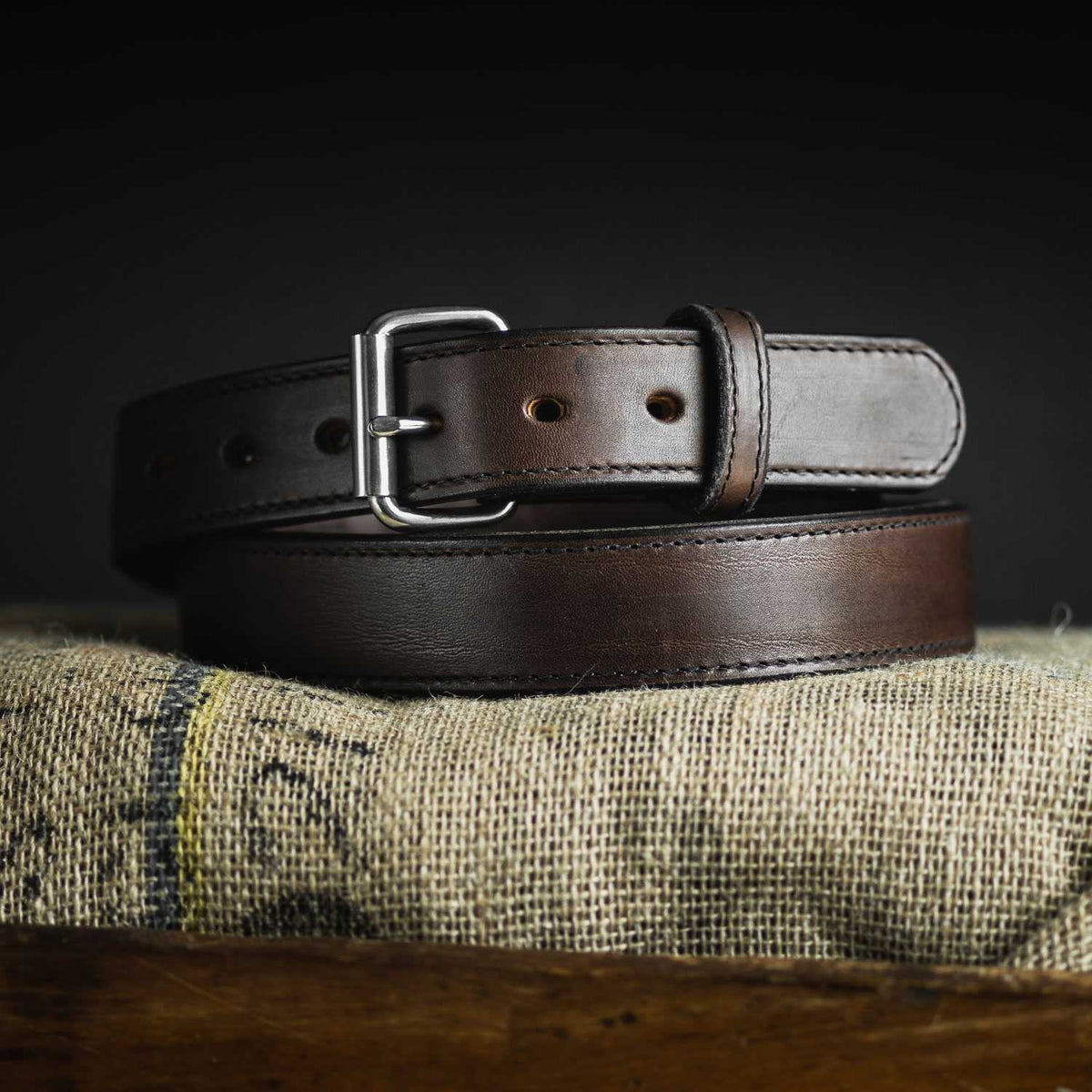 A close-up of a dark brown Hanks Belts Steel Core Belt - 1.5" with a silver buckle, coiled neatly on a piece of burlap fabric. The belt features several punched holes for adjustment. The background is dark, accentuating the rich texture and color of the leather.