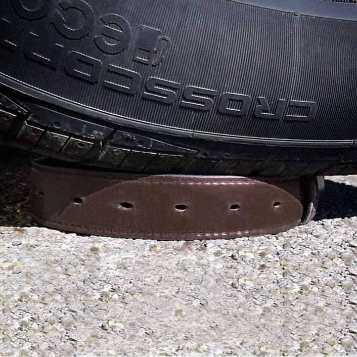 A car tire rests on top of the Hanks Belts Steel Core Belt - 1.5", which lies on a rough concrete surface, visibly flattened by the tire's weight.