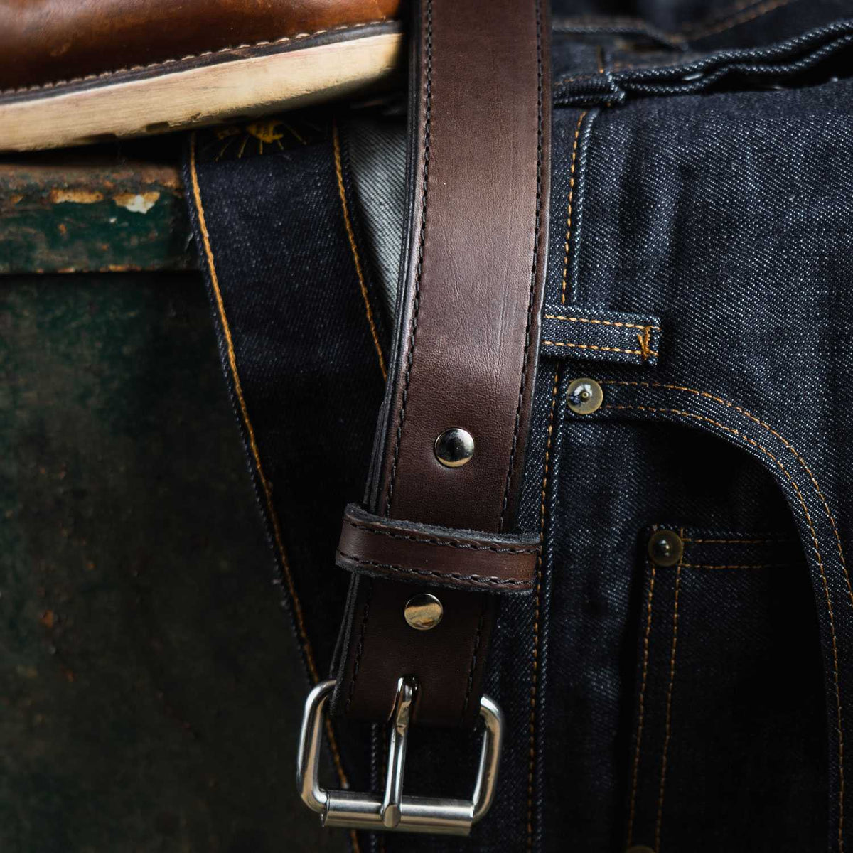 The Steel Core Belt - 1.5" by Hanks Belts, made of dark brown leather and featuring a silver buckle, is draped over a pair of dark blue jeans that have contrast stitching. The belt loop is fastened with a silver snap. Both the jeans and belt rest on a textured green surface, with a brown shoe partially visible above them.