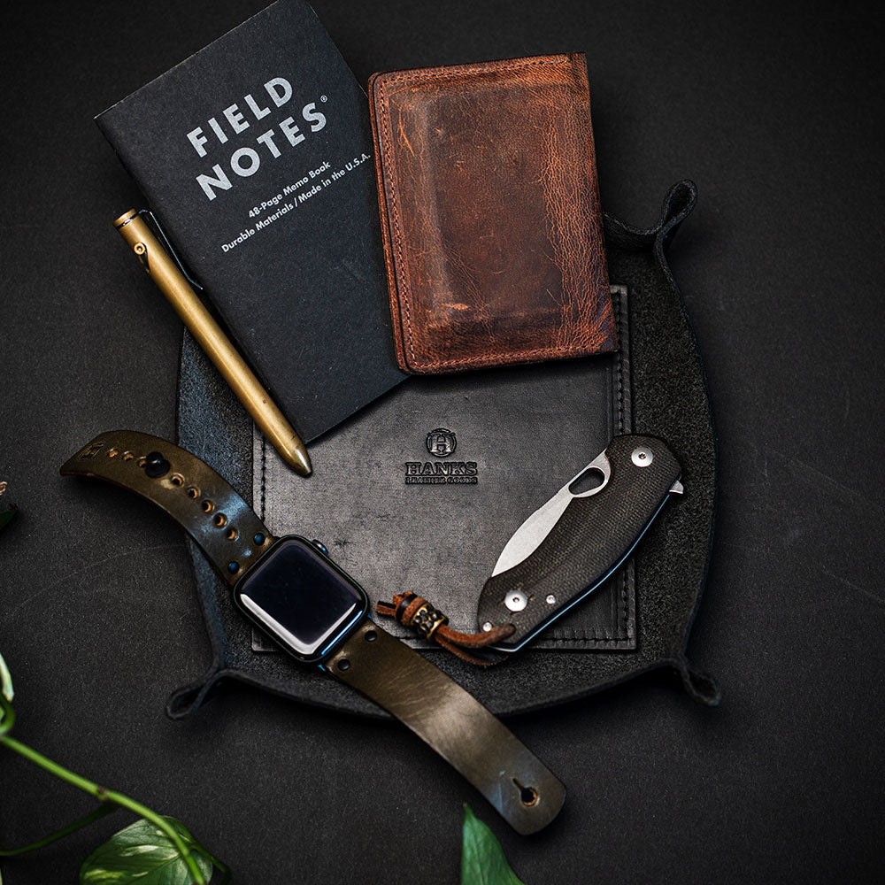 A flat lay photo showcases a leather watch, folding knife, brass pen, leather wallet, and Field Notes notebook on a Hanks Belts Premium Leather Valet Tray. Some green leaves peek in from the bottom left corner, suggesting an assembly of everyday carry essentials.