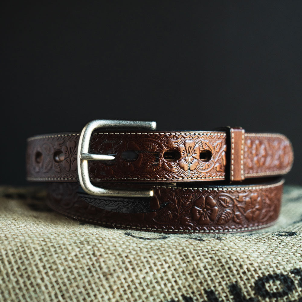 The Floral Embossed by Hanks Belts, a full-grain leather belt with a silver buckle and floral embossing, is coiled on textured beige fabric against a dark background.
