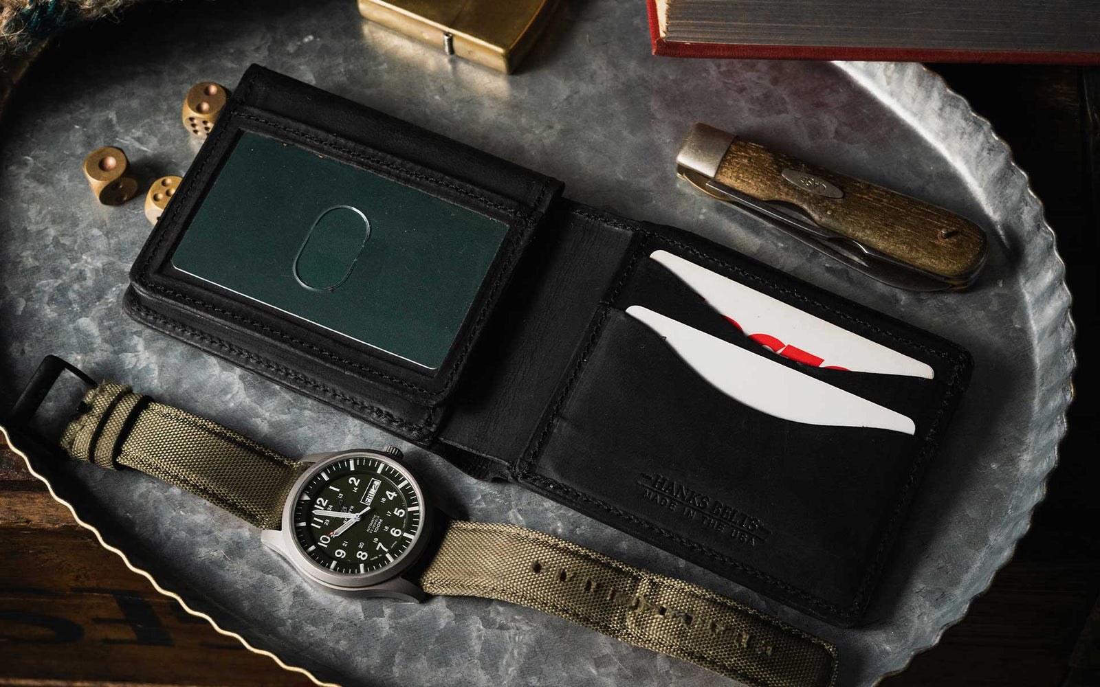 A black leather wallet with cards, a green credit card holder, a pocket knife, a watch with a green strap, and bullets are placed on a round metal tray next to a book.