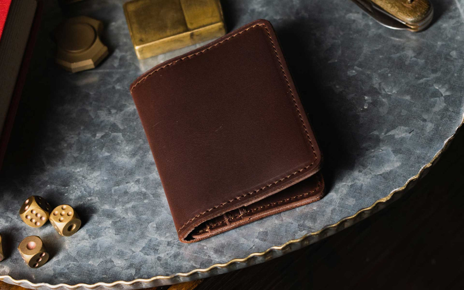 A brown leather wallet is placed on a textured metal surface, surrounded by various objects including dice, a brass lighter, and a small round object.