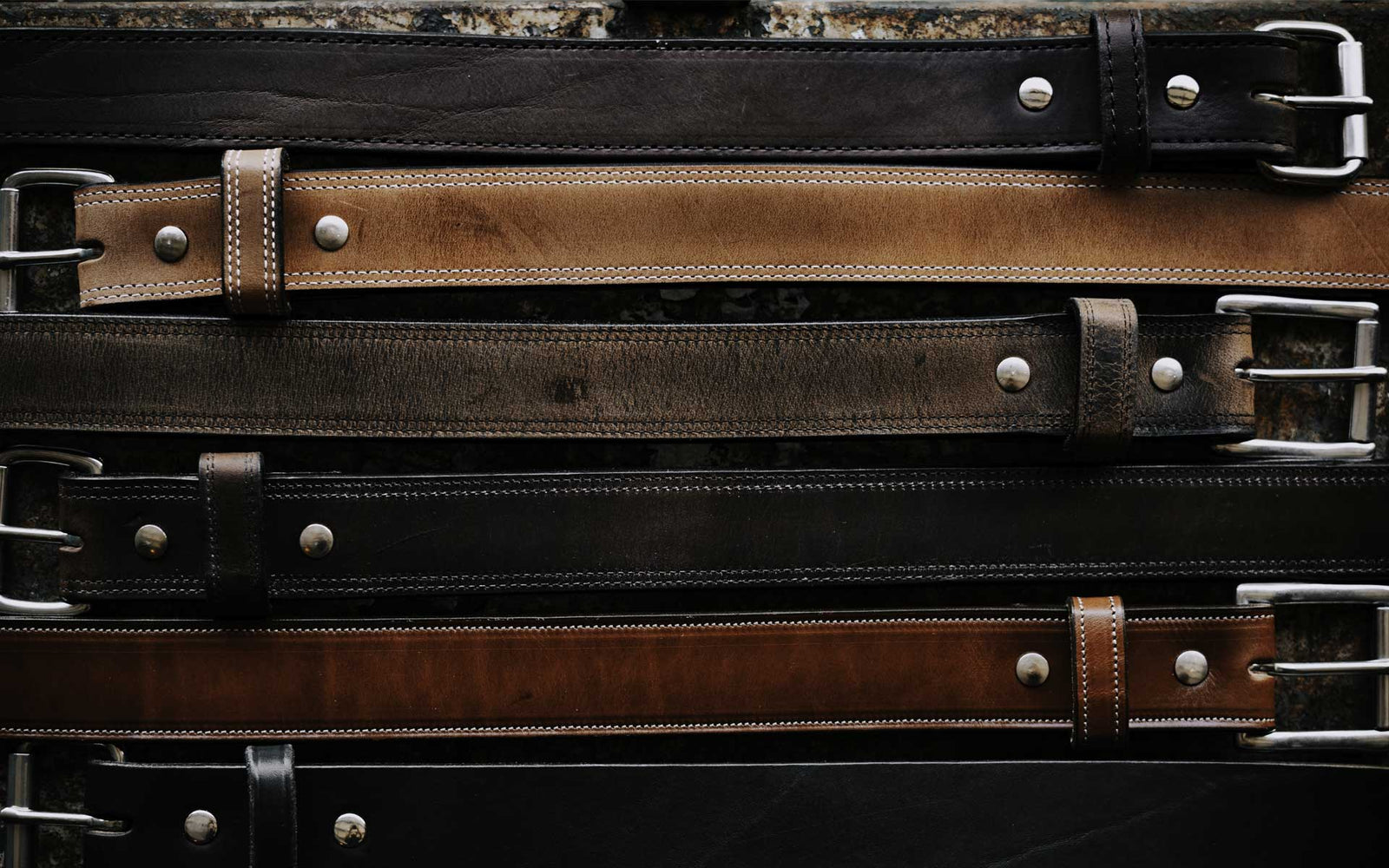 A variety of leather belts in shades of brown and black, displayed vertically. Each belt features a metal buckle and visible stitching, some with metal studs. The belts are arranged on a rustic wooden background.