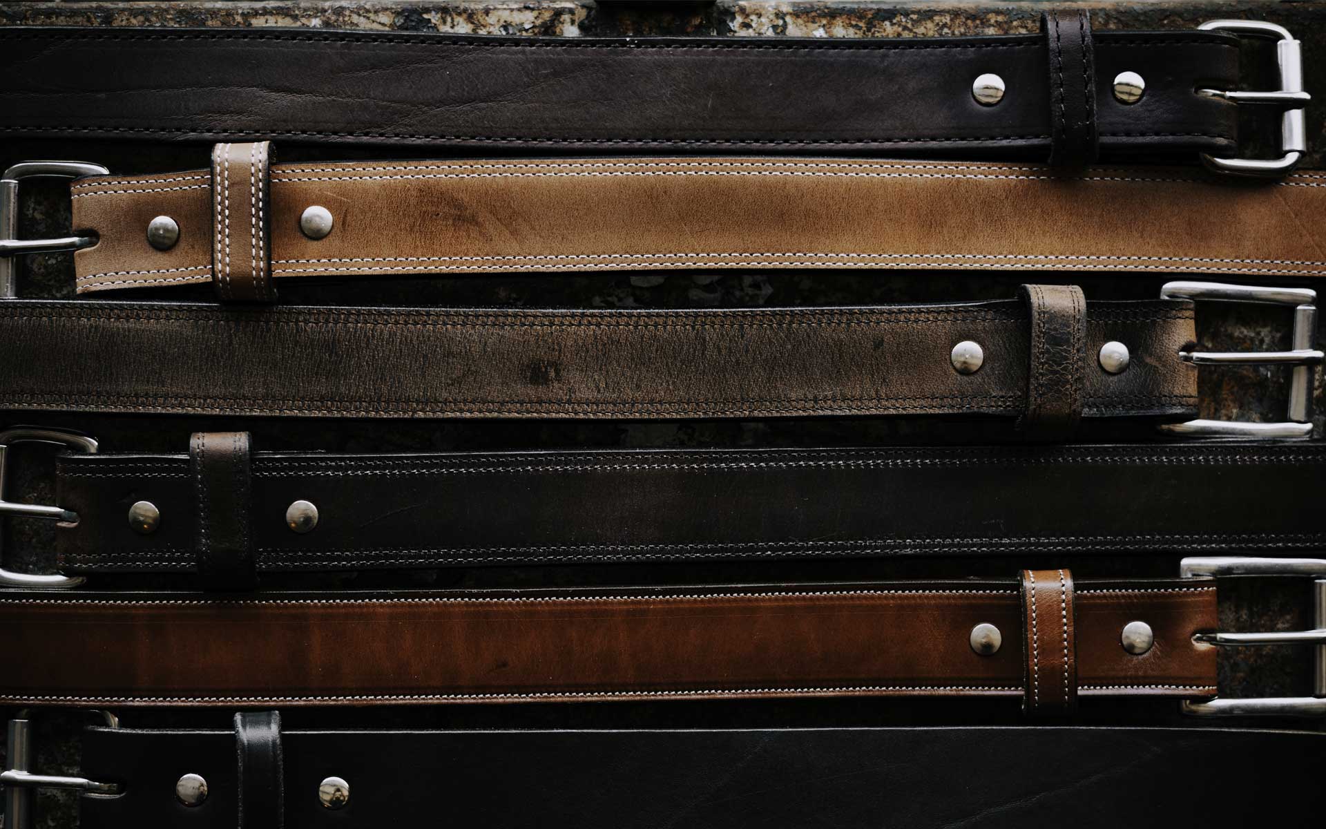 A variety of leather belts in shades of brown and black, displayed vertically. Each belt features a metal buckle and visible stitching, some with metal studs. The belts are arranged on a rustic wooden background.