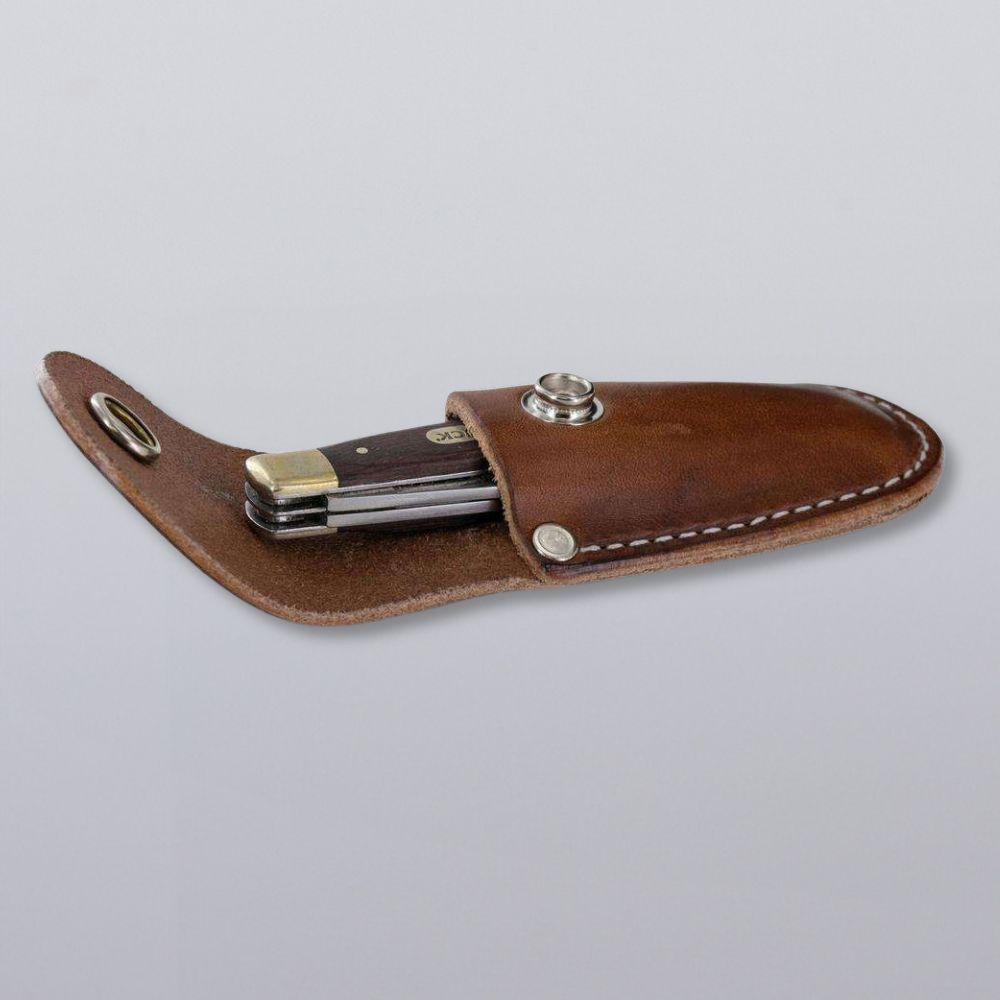 The Hanks Belts Canyon Utility Sheath and Mag Case features a dark-handled folding pocket knife, partially tucked into its USA-made brown leather sheath with white stitching and snap closure, shown on a plain gray background.